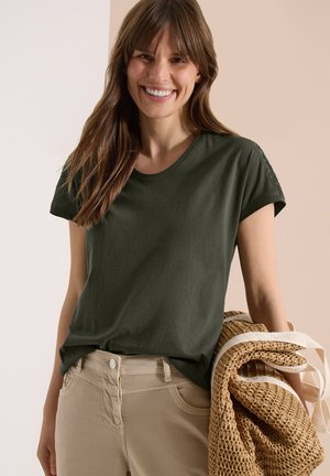 Femme souriante aux longs cheveux bruns portant un t-shirt vert foncé et un pantalon beige, tenant un sac fourre-tout tissé roulé avec des anses blanches.