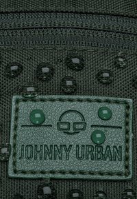 Pezzo di tessuto verde con testi in rilievo "JOHNNY URBAN", contornato da un bordo testurizzato. La superficie presenta gocce lucide e idrorepellenti.