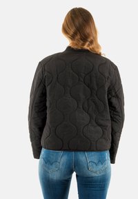 Zwarte gewatteerde jas met een golvend patroon, voorzien van een ronde kraag en lange mouwen. Ggedragen met blauwe slim-fit jeans.