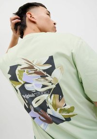 Green cotton T-shirt featuring a large, colorful floral graphic print on the back against a black square with the text "Originals STUDIO."