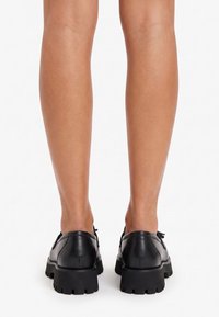 Persona de pie con zapatos de cuero negros de suela gruesa, vista desde atrás que muestra las pantorrillas y los tacones de los zapatos sobre un fondo blanco.