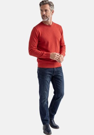 Harmaatukkainen ja -partainen mies, jolla on punainen neulepusero, tummat farkut ja mustat juhlakengät, seisoo kädet ristissä ja katsoo vasemmalle.