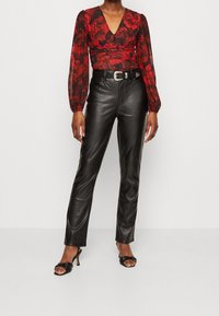Haut à manches longues à motif floral rouge avec un décolleté en V prononcé, associé à un pantalon en cuir noir ajusté et des sandales à talons noires. Ceinture avec boucle en argent.