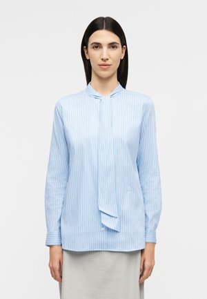 Žena s rovnými tmavými vlasmi, oblečená v svetlomodrej a bielo pruhovanej blúzke s kravatou na krku a svetlosivej sukni, stojaca pred bielym pozadím.