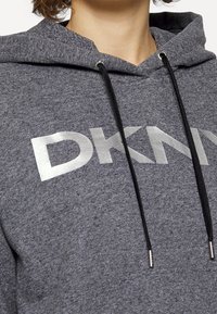 Šedá mikina s kapucí vyrobená z jemné látky, s šedým logem "DKNY", černými šnúrkami a kontrastními stříbrnými detaily.