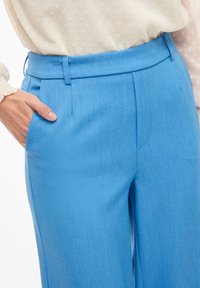 Světlé modré kalhoty s hladkou texturou, dokonale přizpůsobeným designem a bočními kapsami. Mají plochý přední díl a pás pro elegantní vzhled.