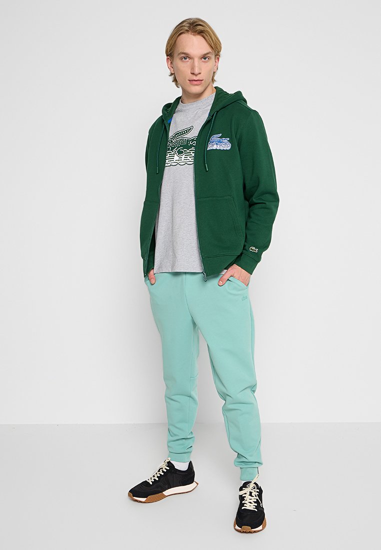 Sweat à fermeture éclair vert avec un design graphique sur la poitrine gauche, associé à un pantalon de jogging mint clair et des baskets noires.