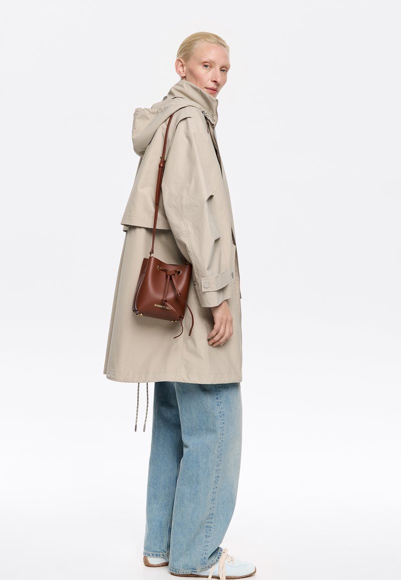 Abrigo impermeable beige con capucha, usado sobre unos vaqueros azules holgados. Incluye una bolsa de cuero marrón con cordón. Diseño minimalista y colores neutros.