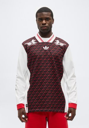 Άνδρας που φοράει γιλέκο με σχέδιο AFC σε μαύρο και κόκκινο πάνω από λευκό πουκάμισο με μακριά μανίκια, κόκκινες μανσέτες και κόκκινο παντελόνι, αντιμετωπίζοντας μπροστά.