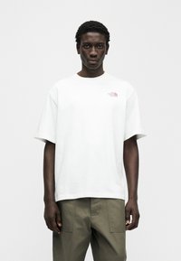 Λευκό βαμβακερό μπλουζάκι με κοντά μανίκια και μικρό ροζ λογότυπο στο στήθος. Συνδυασμένο με χακί παντελόνι. Απλό φόντο.