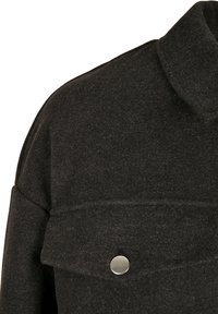 Épaule de veste en laine gris foncé avec une poche à rabat sécurisée par un seul bouton-pression rond en argent.