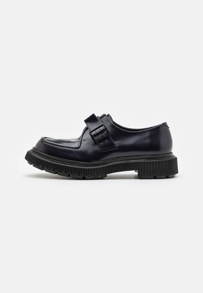 Pantofi din piele patent neagră, cu vârf rotund, două curele reglabile și o talpă groasă, cu caneluri. Textură netedă, cu detalii minime.