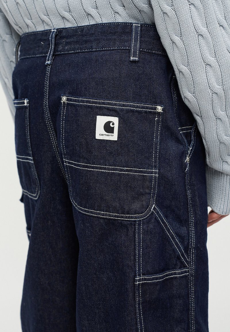 Gros plan sur une personne portant un jean Carhartt bleu foncé avec des coutures blanches visibles et un pull en maille torsadée gris.