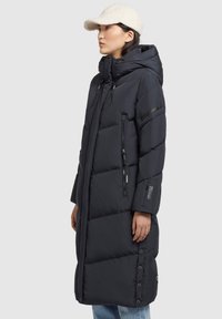Cappotto puffo nero con cappuccio, tasche con zip e bottoni a pressione. Presenta una texture trapuntata e un cordino regolabile. Indossato con un berretto color crema.
