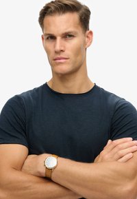 Reloj con correa de cuero marrón claro, con una esfera plateada redonda y detalles dorados, llevado en un brazo desnudo. La persona lleva una camiseta de color azul oscuro.