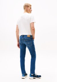 Młody mężczyzna w białej koszulce i niebieskich dżinsach stoi z rękami w kieszeniach, zwrócony tyłem i patrzący na bok na jednolitym białym tle.