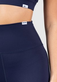 Granatowe legginsy wysokiej talii, wykonane z gładkiego, elastycznego materiału, z płaskimi szwami i małą białą metką z logo na pasie.