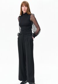 Zwarte geribbelde top met doorschijnende zwarte mouwtjes met stippen, gecombineerd met zwarte broek met hoge taille voorzien van plooien en zijzakken.