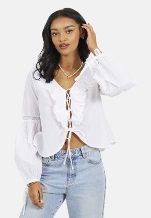 Kobieta w białej bluzce z falbankami, z ozdobnym wiązaniem z przodu i bufkami, w połączeniu z jasnoniebieskimi dżinsami, dotykająca włosów i zwrócona przodem.