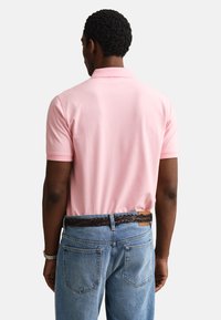 Muž nosící světle růžové polo triko s krátkým rukávem zastrčené do modrých džín s tmavým pleteným opaskem, viděn zezadu.