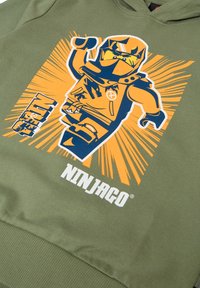 Grön hoodie med en stor orange och blå LEGO Ninjago-grafik av en ninja, med fet text och burst-designelement. Mjuk tygtextur.