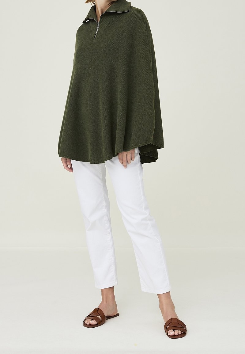 Maglione oversize verde oliva con collo a zip, lavorazione a maglia strutturata, abbinato a pantaloni bianchi a gamba dritta e sandali bassi marroni.