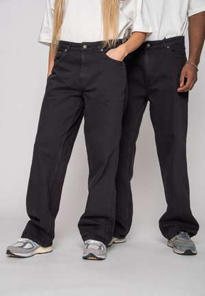 Zwei Personen tragen schwarze, locker sitzende Jeans und weiße T-Shirts, stehen nebeneinander und haben ihre Hände in oder nahe den Taschen.