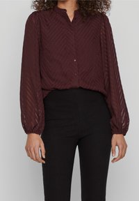 Blouse bordeaux avec un motif en chevrons, manches longues en voile, col montant et patte de boutonnage, associée à un pantalon noir taille haute.