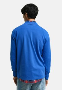 Blauwe polo met lange mouwen van een gladde stof met een kraag. Draagt over een rood-blauw geruit overhemd, gecombineerd met lichtdenim jeans.