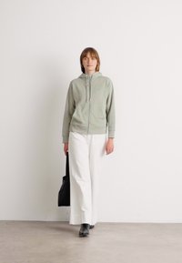 Felpa con zip color verde menta in un tessuto testurizzato, abbinata a pantaloni larghi bianchi e stivali neri, insieme a una borsa tote nera.