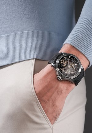 Vīrieša roka bēšā bikšu kabatā, nēsājot melnu gumijas siksniņu pulkstenim ar redzamu mehānisko skeleta ciparnīcu un sudraba rāmi.