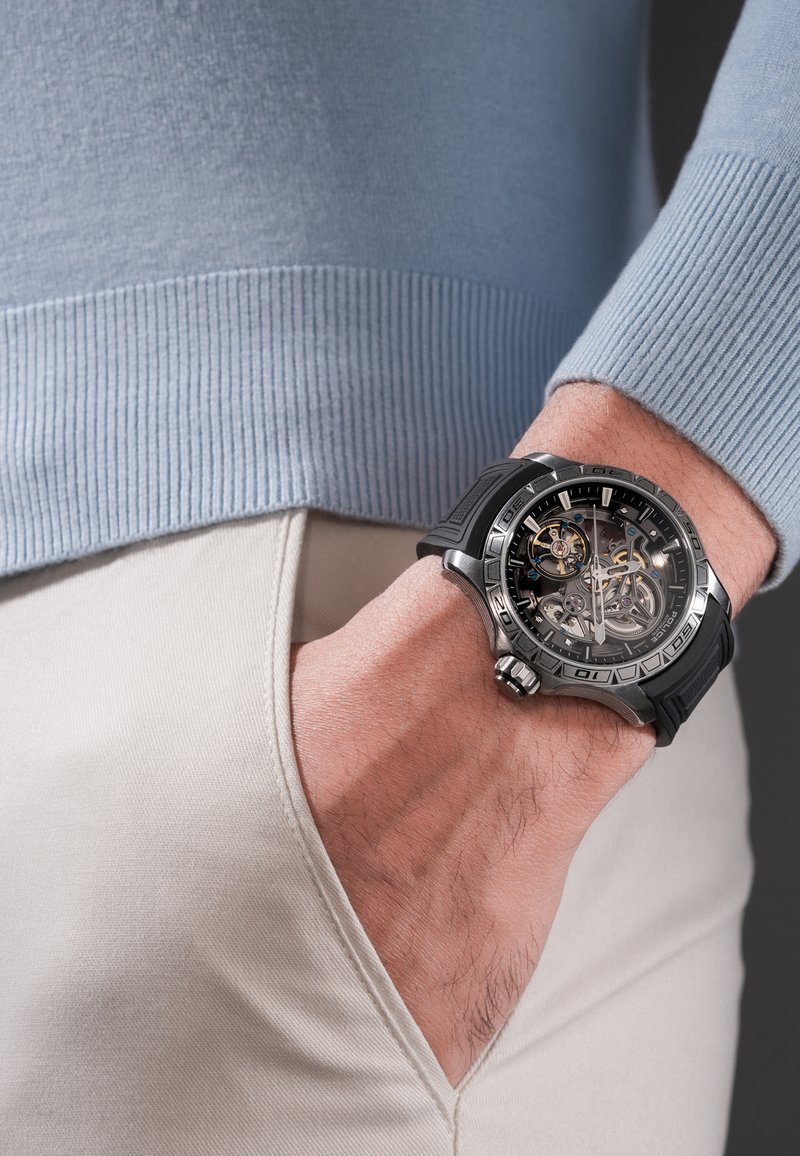 Mano masculina en el bolsillo de pantalones beige, con un reloj de pulsera de correa de goma negra, esfera esqueleto mecánica visible y bisel plateado.