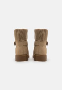 Tanngule støvler med en strikket tan mansjett. Sueskinns tekstur, bakre spenne detalj, og en brun gummisåle. Minimalistisk design med en nøytral palett.