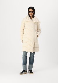Krémový puff coat s širokým golierom a vzorom v tvare chevronu, kombinovaný s roztrhnutými modrými džínsami a čiernymi teniskami s bielymi pruhmi.