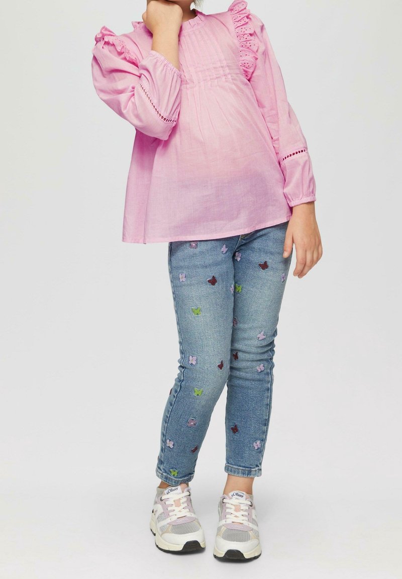 Enfant portant un chemisier rose à manches longues avec des volants, un jean bleu avec un motif papillon et des baskets blanches, debout avec une main près du visage.