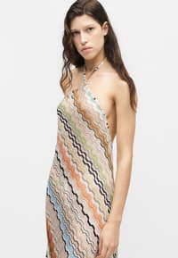 Joven mujer con cabello largo y castaño que lleva un vestido sin mangas con tirantes halter con patrones ondulados multicolores en zigzag sobre fondo blanco.