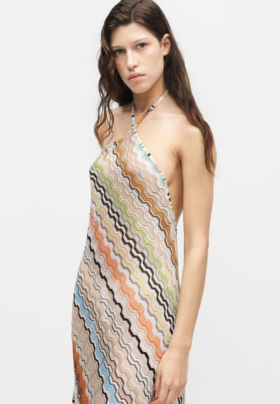Jeune femme aux longs cheveux bruns portant une robe dos nu sans manches avec des motifs ondulés en zigzag multicolores sur fond blanc.