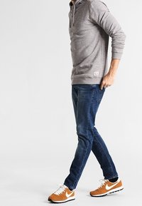 Szary hoodie o teksturowanym wyglądzie, niebieskie dżinsy oraz pomarańczowo-białe trampki. Stylizacja ma swobodny, sportowy charakter.