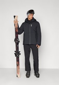 Schwarze isolierte Skijacke, schwarze Hose und schwarze Skischuhe. Hält Skier mit Bindungen. Graue Strickmütze und Skibrille.