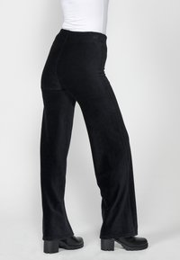 Personne portant un pantalon noir taille haute à jambes larges et des bottes noires à talons épais sur un fond clair uni.