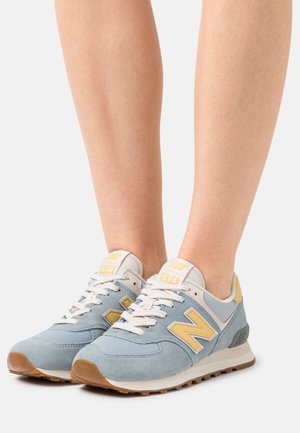 Pieds chaussés de baskets New Balance 574 bleu clair et jaune avec des lacets blancs et des semelles couleur gomme sur fond blanc.