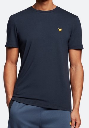 Mężczyzna ubrany w dopasowaną, granatową koszulkę z krótkim rękawem z małym żółtym logo ptaka na lewej piersi.