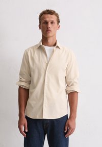 Beżowa koszula zapinana na guziki z podwiniętymi rękawami, wykonana z lekkiego materiału; ma klasyczny kołnierzyk i prosty dół. Noszona na białym T-shircie.
