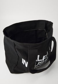 Bolsa de tela negra con una amplia apertura, fabricada con un material resistente, asas cortas y un bolsillo con cremallera en el interior. Presenta texto en blanco destacado en el lateral.