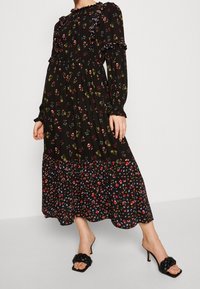 Abito nero floreale con maniche lunghe, scollatura a volant e gonna a livelli. Presenta motivi floreali multicolori ed è abbinato a mule con tacco nere.