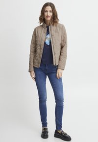 Jachetă bej matlasată cu guler înălțat, asociată cu un tricou grafic bleu marin, blugi skinny albaștri și mocasini negri. Textură netedă, design simplu.