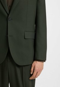 Mörkgrön skräddarsydd kavaj med en slät yta, enfaldig design, två knappar och en bröstfickdetalj.