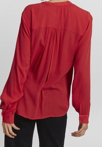 Blusa roja texturizada con mangas largas y puños abotonados. Presenta un yugo trasero fruncido y un dobladillo redondeado.