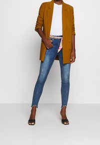 Hořčicový blazer, bílý tričko, modré skinny džíny s třepeným lemem a černé otevřené lodičky. Džíny mají jemné zdobení.