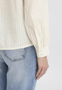 Blusa crema con sottili righe verticali, maniche lunghe, polsini con bottoni, realizzata in tessuto leggero e con una vestibilità morbida, abbinata a jeans azzurri chiari.